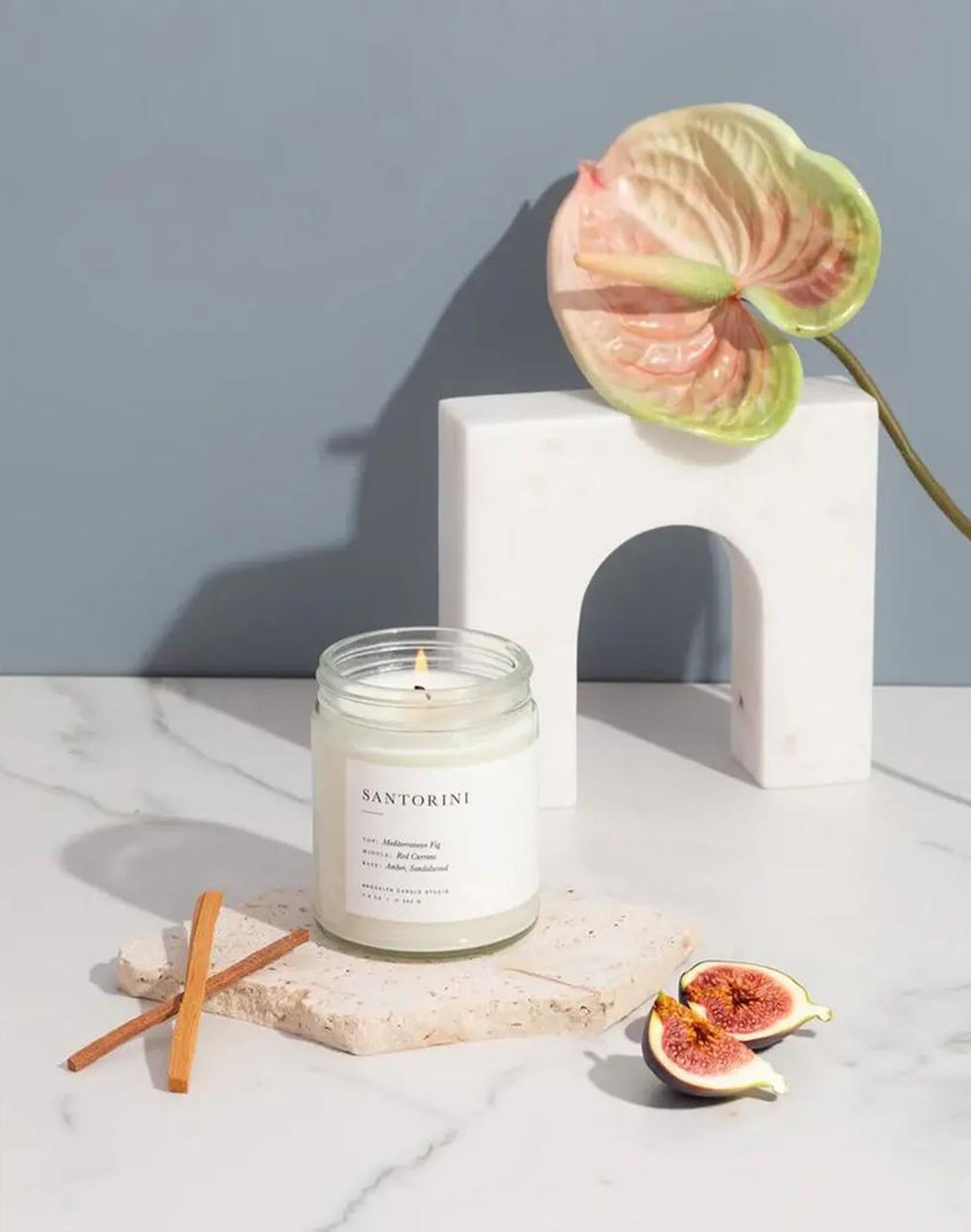 The Brooklyn Candle Studio Santorini Jar Candle, scented with Mediterranean Fig, rests on a stone coaster with a cinnamon stick and halved fig. A white arch sculpture and pink-green anthurium flower evoke serene Greek Isles vibes against a gray background.