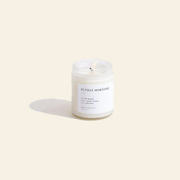 A lit Brooklyn Candle Studio Sunday Morning Jar Candle sits on a light beige background, its glass jar label visible. The candle releases soothing jasmine and amber woods notes, casting a soft shadow to the left.