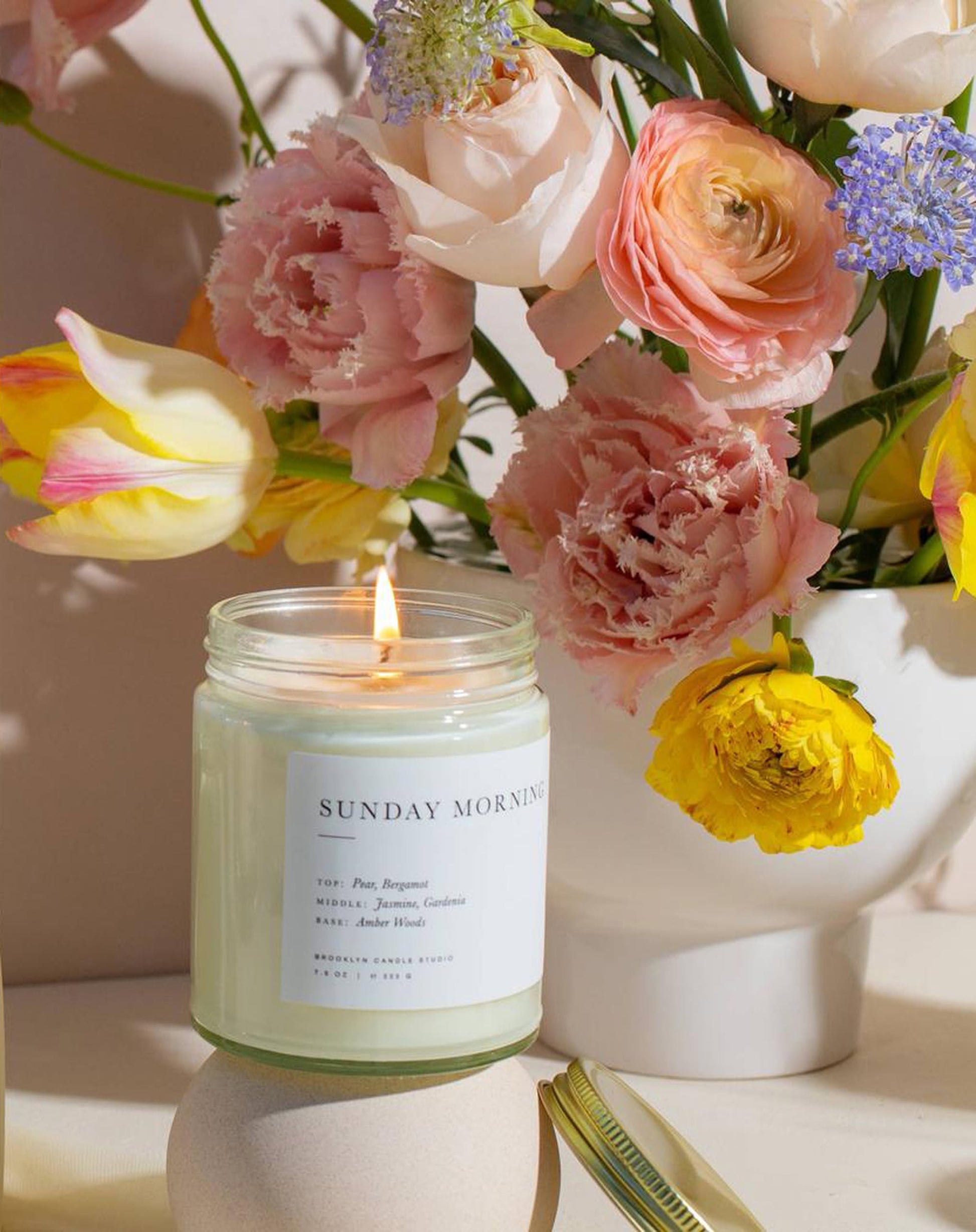 A Brooklyn Candle Studio Sunday Morning Jar Candle glows beside a white vase of colorful flowers, filling the space with calming amber woods notes for a fresh, inviting atmosphere.