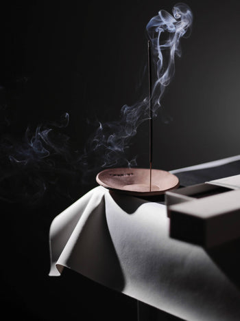 A CHANIDA Hinoki-Rose Incense Stick from CHANIDA stands lit in a round holder on a draped white cloth, its smoke curling upward against a dark background, with the products rectangular box placed nearby on the table.