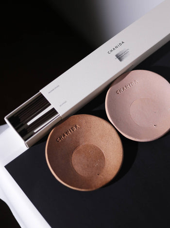 A white CHANIDA box of Japanese Green Tea-Vanilla Incense Sticks sits beside two round brown ceramic holders stamped CHANIDA, all arranged on a black and white surface, evoking an elegant Japanese ambiance.
