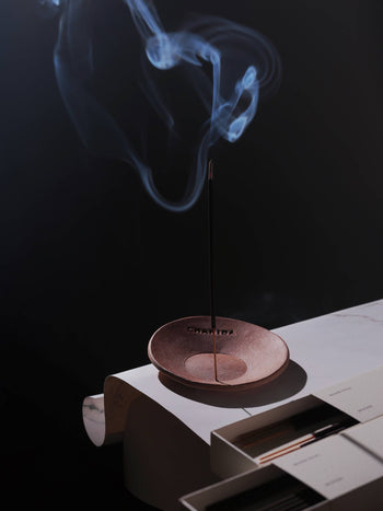 A CHANIDA Lavender-Musk Incense Stick burns in a small dish on a white surface beside a closed book, releasing curling smoke against a dark background.