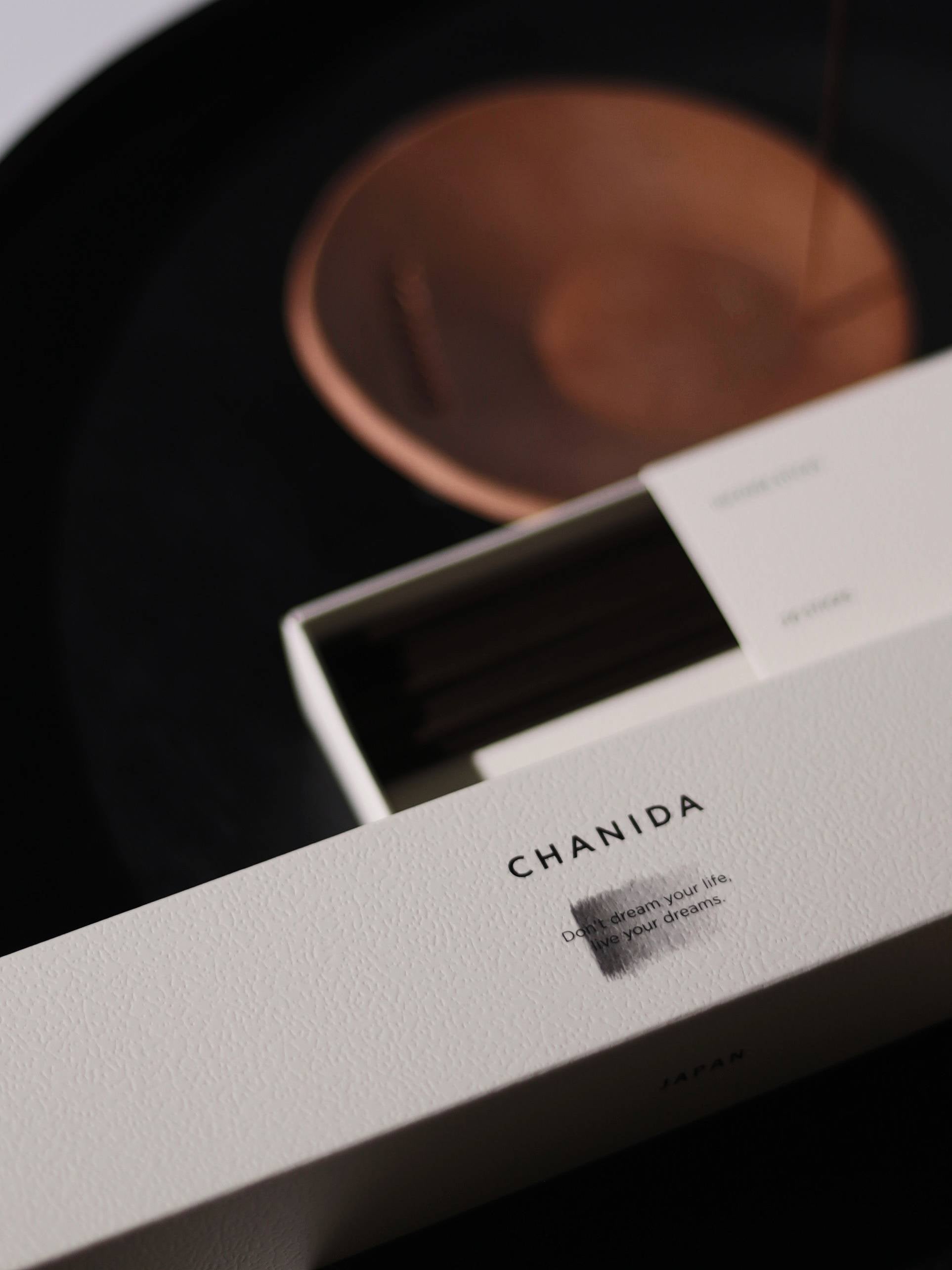 A close-up of a partially opened CHANIDA Lignum Juniperi-Cypress Incense Sticks box reveals dark incense sticks inside with a juniper wood scent, while a blurred copper holder sits in the background on a dark surface.