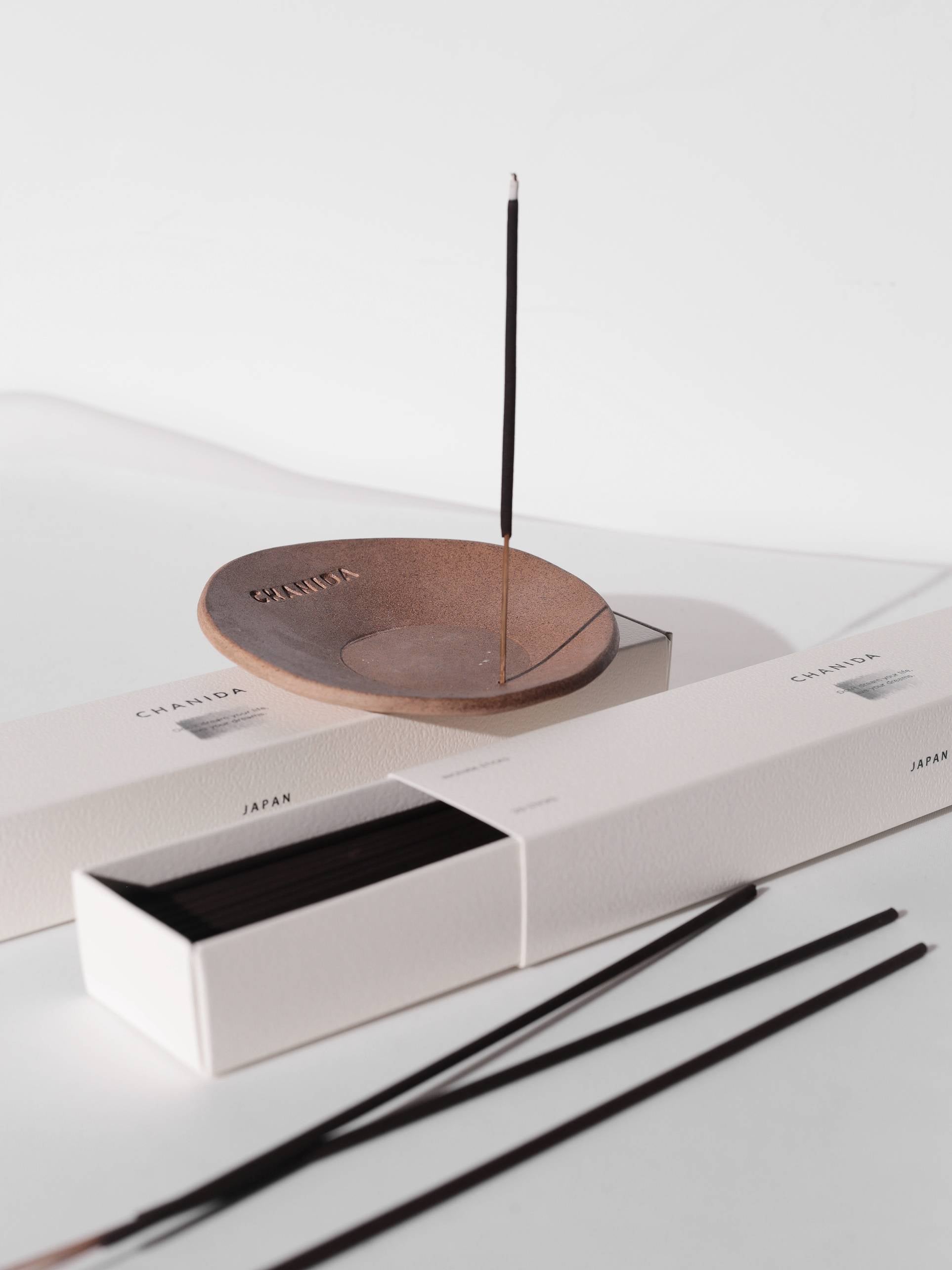 A lit CHANIDA Lignum Juniperi-Cypress incense stick stands upright in a brown ceramic holder, releasing a soothing fragrance beside white CHANIDA boxes containing more incense sticks.