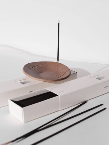 A lit CHANIDA Lignum Juniperi-Cypress incense stick stands upright in a brown ceramic holder, releasing a soothing fragrance beside white CHANIDA boxes containing more incense sticks.