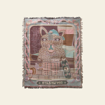 The DETAIL Black Cat Oax Throwket by DETAIL features a stylized black cartoon cat with brown stripes, wearing a beanie, holding a skateboard on brickwork. This multicolour throw blanket has pastel borders and “Soy Un Gato” at the bottom.