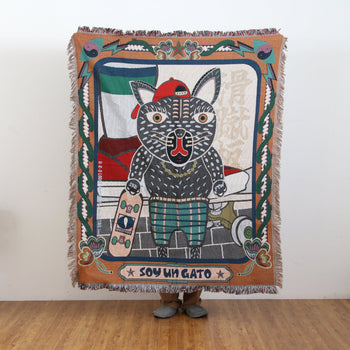 A person is holding the DETAIL Black Cat Oax Throwket by DETAIL, a vibrant multicolour blanket featuring a cartoon cat in a red cap with a skateboard, SOY UN GATO at the bottom, and decorative border patterns.