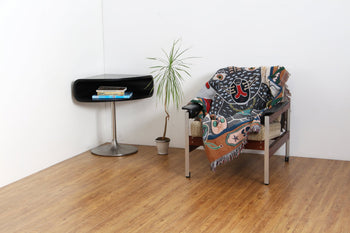 A modern, minimalist room features a chair draped with the DETAIL Black Cat Oax Throwket by DETAIL, a small potted plant, and a black side table with books on wooden floors against white walls.