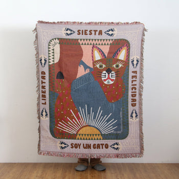 A person holds up the DETAIL Cat Oax Throwket by DETAIL, a vibrant throw blanket featuring colorful folk art cat motifs, Spanish words like “Siesta” and “Libertad,” and abstract shapes—only their feet are visible behind it.
