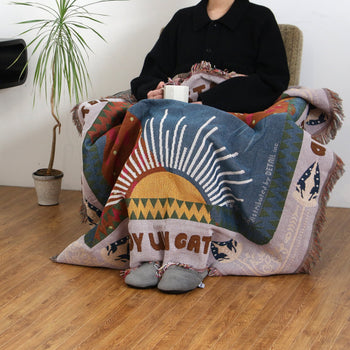 A person sits on a chair holding a white mug, wrapped in the DETAIL Cat Oax Throwket by DETAIL featuring sun and cat designs. The wooden floor and a nearby potted plant complete the scene, with their face not shown.
