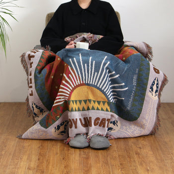 A person in a black coat sits on a chair with the DETAIL Cat Oax Throwket by DETAIL, featuring a sun design, draped over their lap while holding a white mug. The floor is wooden and theres a plant visible on the left.