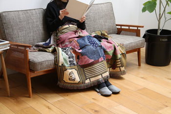 Wrapped in the vibrant DETAIL Dodo Oax Throwket by DETAIL, a person wearing dark clothes and gray slippers relaxes on a sofa reading, surrounded by wooden floors, a potted plant, and Oaxaca-inspired décor in a cozy living room.