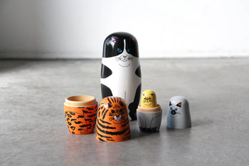 The DETAIL Feline 5 Nesting Dolls by DETAIL feature wooden dolls in various sizes, each painted to resemble a different cat. Arranged on a gray surface, these nesting dolls are perfect for cat lovers with their unique expressions and patterns.