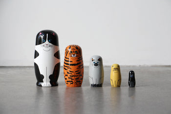The DETAIL Feline 5 Nesting Dolls by DETAIL features five wooden cat-shaped dolls of different sizes and colors, arranged on a smooth gray floor against a white wall. Each doll is painted to resemble a unique type of cat—perfect for cat lovers.