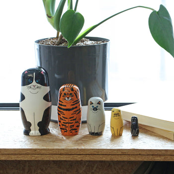 The DETAIL Feline 5 Nesting Dolls by DETAIL, in varying sizes, are arranged on wood near a gray potted plant and an open book with natural light—charming decor for cat lovers.