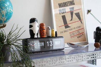 A metal shelf displays a flip clock set to “Sun 13 May,” the DETAIL Feline 5 Nesting Dolls by DETAIL, a globe, a modern spoon sculpture, a plant, and a framed marketing poster leaning against the wall.