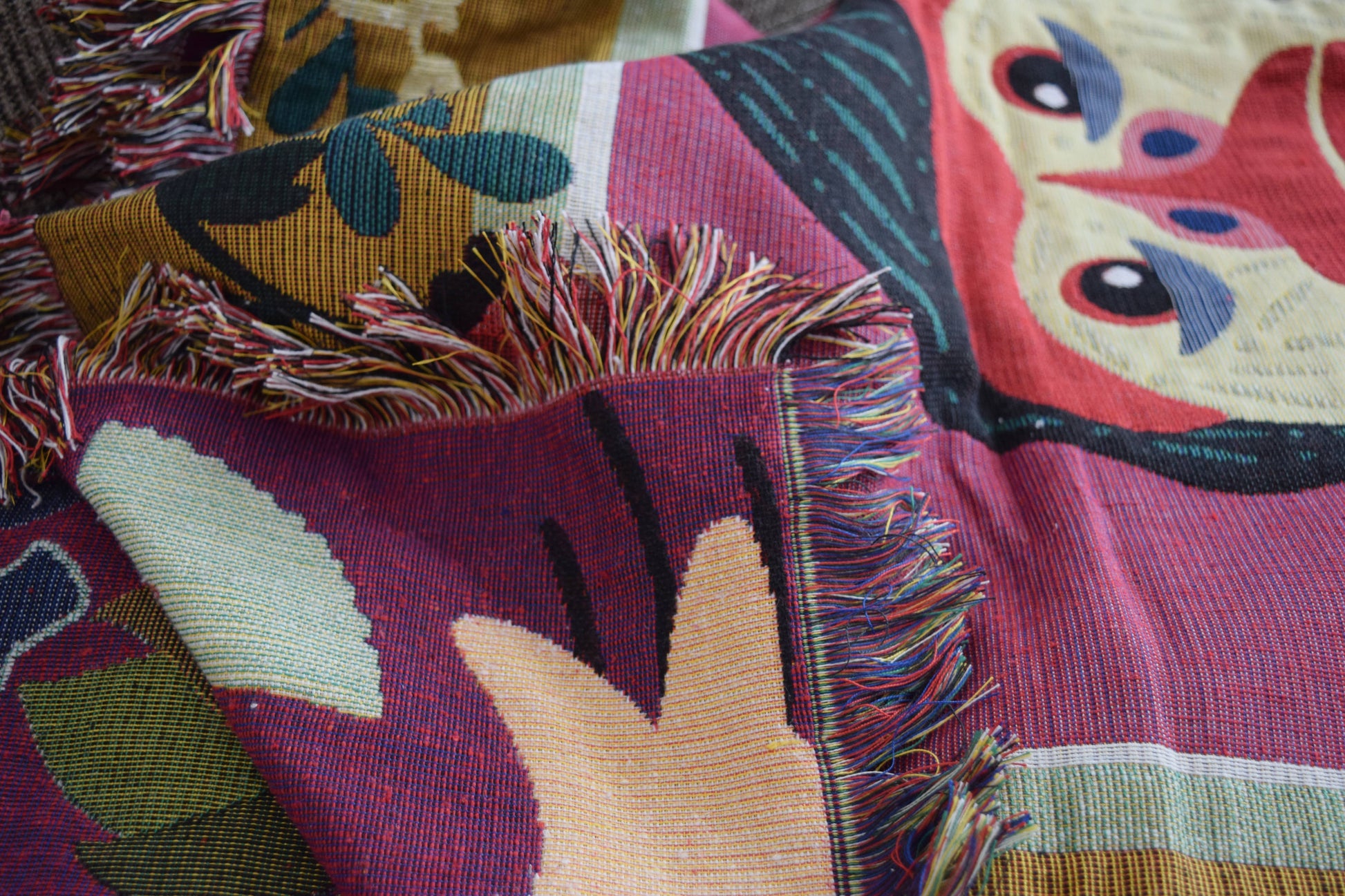 A close-up of the DETAIL Gorilla Oax Throwket by DETAIL, a multicolour throw blanket inspired by Oaxaca folk art, featuring abstract shapes, leaves, a whimsical face, and bright fringe in red, green, cream, and yellow.