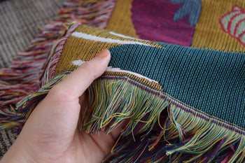 A hand holds the fringed edge of the DETAIL Gorilla Oax Throwket by DETAIL, displaying its colorful woven fabric with green, yellow, and multicolored threads and floral patterns—a stunning example of Oaxacan folk art.