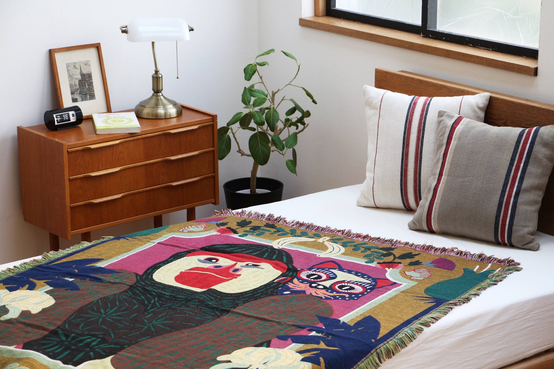 A neatly made bed with two striped pillows and the DETAIL Gorilla Oax Throwket by DETAIL, featuring abstract faces inspired by Oaxaca folk art. Nearby is a wooden nightstand with a lamp, framed photo, book, alarm clock, and a potted plant.