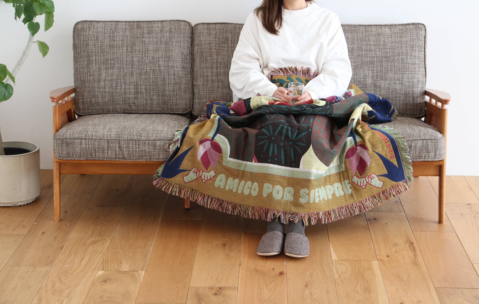 A woman wearing a white shirt and gray slippers sits on a gray sofa with the DETAIL Gorilla Oax Throwket by DETAIL, inspired by Oaxacan folk art, draped over her lap. A potted plant is nearby on the wooden floor.
