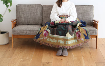 A woman wearing a white shirt and gray slippers sits on a gray sofa with the DETAIL Gorilla Oax Throwket by DETAIL, inspired by Oaxacan folk art, draped over her lap. A potted plant is nearby on the wooden floor.
