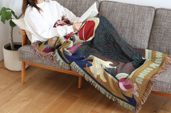 A person relaxes on a gray sofa, reading under the multicolour DETAIL Gorilla Oax Throwket by DETAIL, inspired by Oaxaca folk art. A potted plant sits nearby, and warm wooden flooring completes the cozy scene.