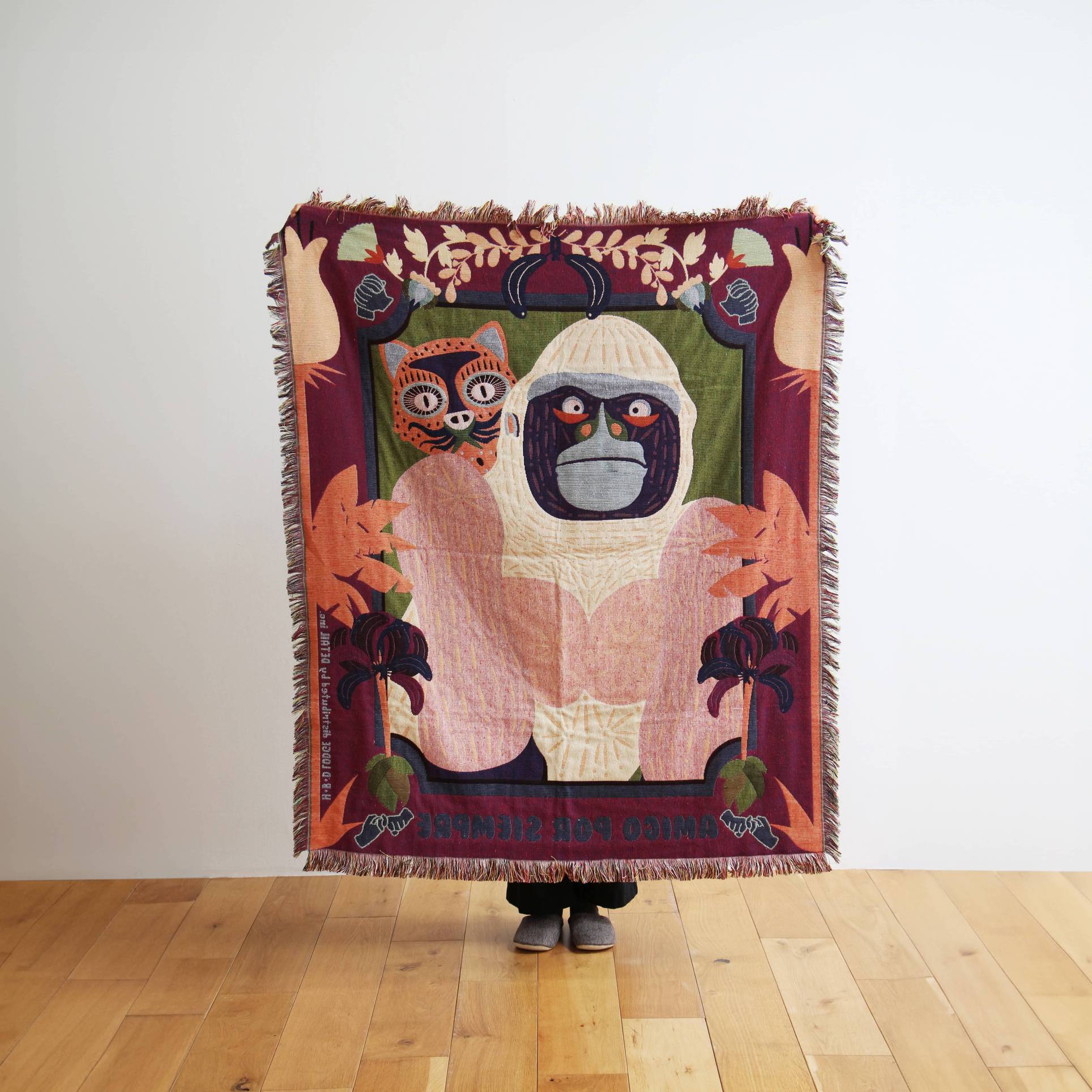 A person holds the DETAIL Gorilla Oax Throwket by DETAIL, a vibrant multicolour blanket inspired by Oaxacan folk art, with a white gorilla and red-orange cat among plants and stars, standing on a hardwood floor against a plain white wall.
