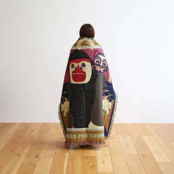 A person faces away, wrapped in the DETAIL Gorilla Oax Throwket by DETAIL, a multicolored blanket inspired by Oaxaca folk art with a bold monkey, pink owl, and foliage design, on light wood flooring against a plain white wall.