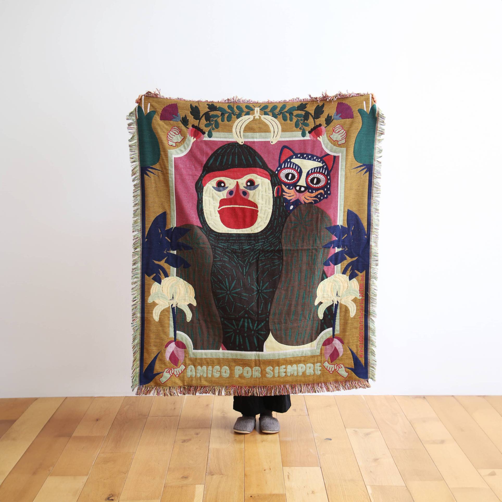 A person holds up the DETAIL Gorilla Oax Throwket by DETAIL, a multicolour throw blanket with cartoonish sloth and cat, leaves, and birds in Oaxaca folk art style. “AMIGO POR SIEMPRE” is at the bottom; only their feet are visible.