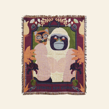 The DETAIL Gorilla Oax Throwket by DETAIL is a woven multicolour throw blanket with fringed edges, featuring a stylized white gorilla with a stern expression, Oaxaca folk art leaf motifs, a small cat-like face, and text at the bottom.