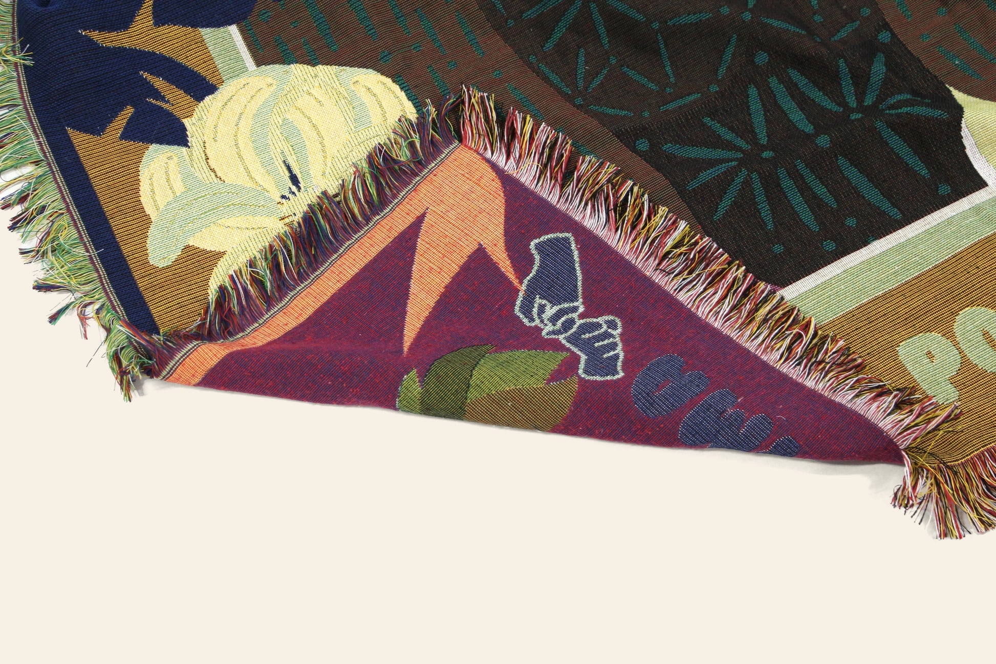 The DETAIL Gorilla Oax Throwket by DETAIL features multicolored woven floral and abstract patterns inspired by Oaxacan folk art, with part of the blanket folded to reveal intricate underside designs and text.