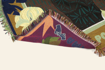 The DETAIL Gorilla Oax Throwket by DETAIL features multicolored woven floral and abstract patterns inspired by Oaxacan folk art, with part of the blanket folded to reveal intricate underside designs and text.