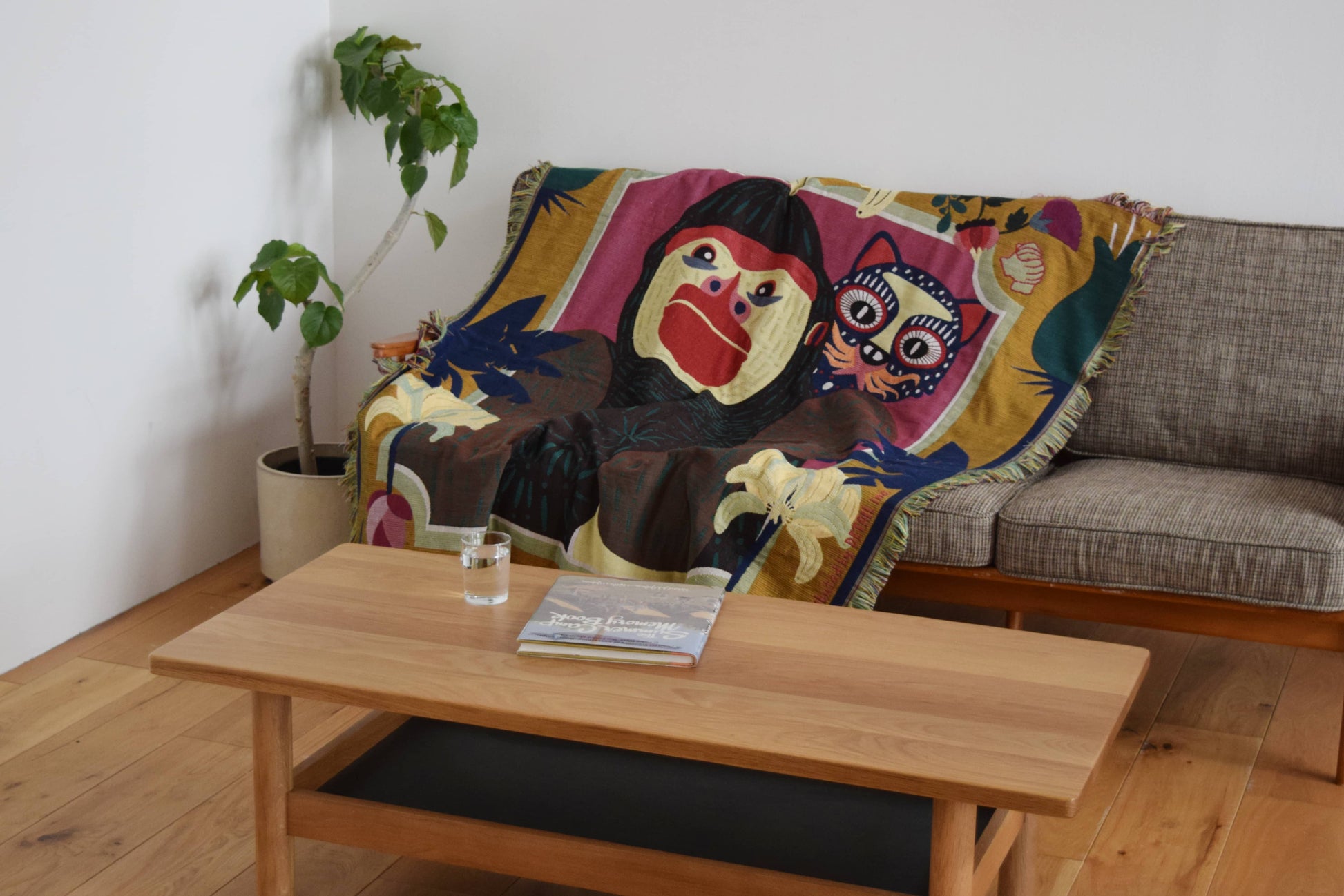 A cozy living room features a gray sofa with the DETAIL Gorilla Oax Throwket—a multicolour throw by DETAIL inspired by Oaxacan folk art, with playful monkey and cat motifs. A plant stands beside a wooden coffee table holding a book and glass of water.