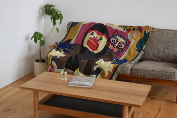 A cozy living room features a gray sofa with the DETAIL Gorilla Oax Throwket—a multicolour throw by DETAIL inspired by Oaxacan folk art, with playful monkey and cat motifs. A plant stands beside a wooden coffee table holding a book and glass of water.