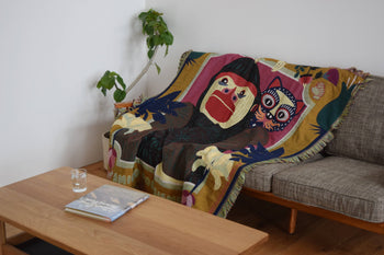 A sofa is draped with the DETAIL Gorilla Oax Throwket by DETAIL, inspired by Oaxaca folk art and featuring a bold monkey and owl design, next to a green plant. In front, a wooden coffee table holds a glass of water and a book.
