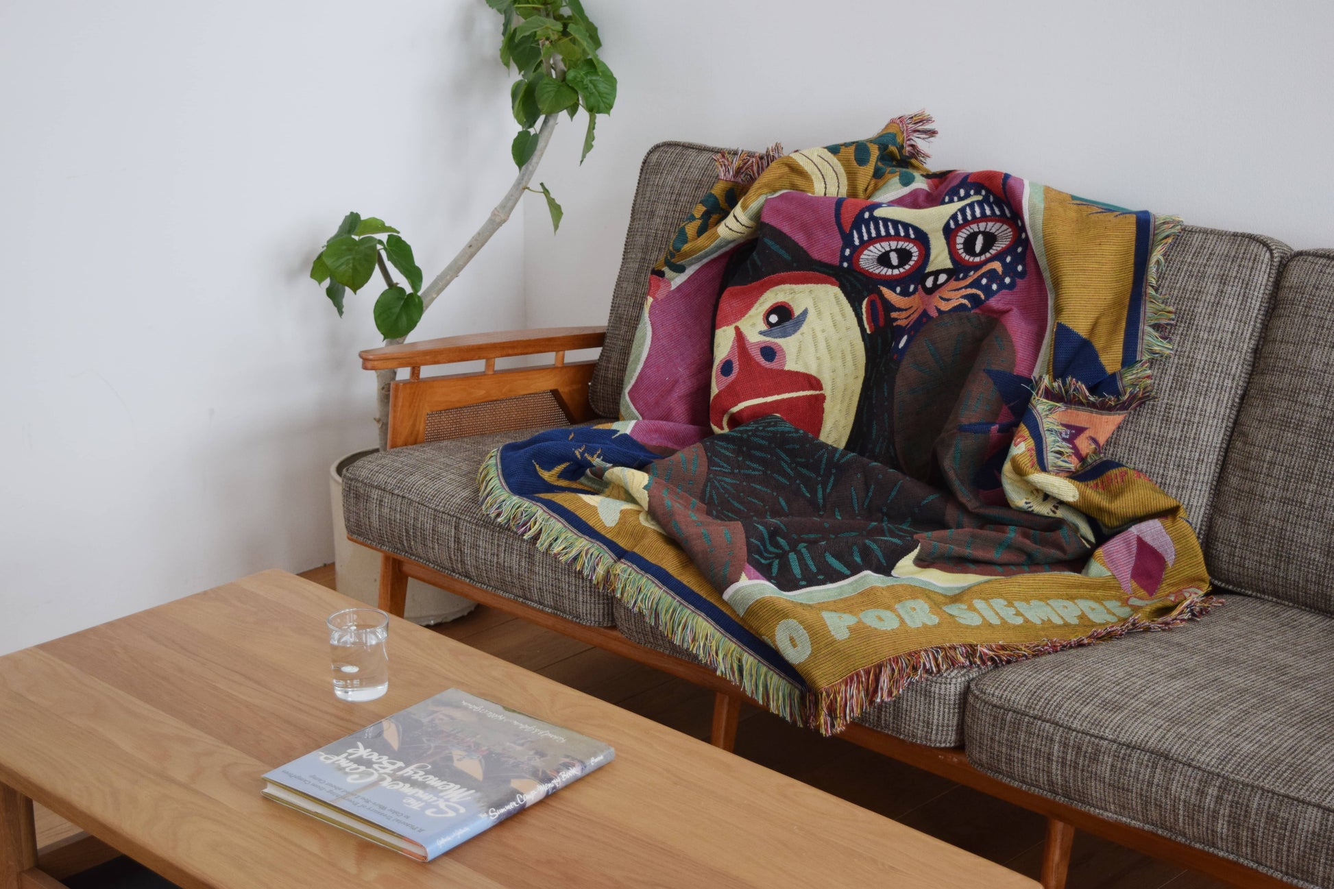 The DETAIL Gorilla Oax Throwket by DETAIL, featuring multicolour abstract faces inspired by Oaxacan folk art, is draped over a gray sofa beside a leafy plant. A book and glass of water sit on the wooden coffee table in this bright, minimal space.