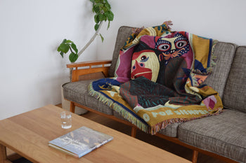 The DETAIL Gorilla Oax Throwket by DETAIL, featuring multicolour abstract faces inspired by Oaxacan folk art, is draped over a gray sofa beside a leafy plant. A book and glass of water sit on the wooden coffee table in this bright, minimal space.