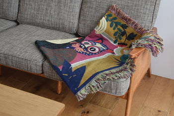 The DETAIL Gorilla Oax Throwket by DETAIL, inspired by Oaxacan folk art and featuring a stylized owl, is draped over the arm and seat of a gray upholstered sofa with wooden arms, beside a light wood coffee table on a wooden floor.