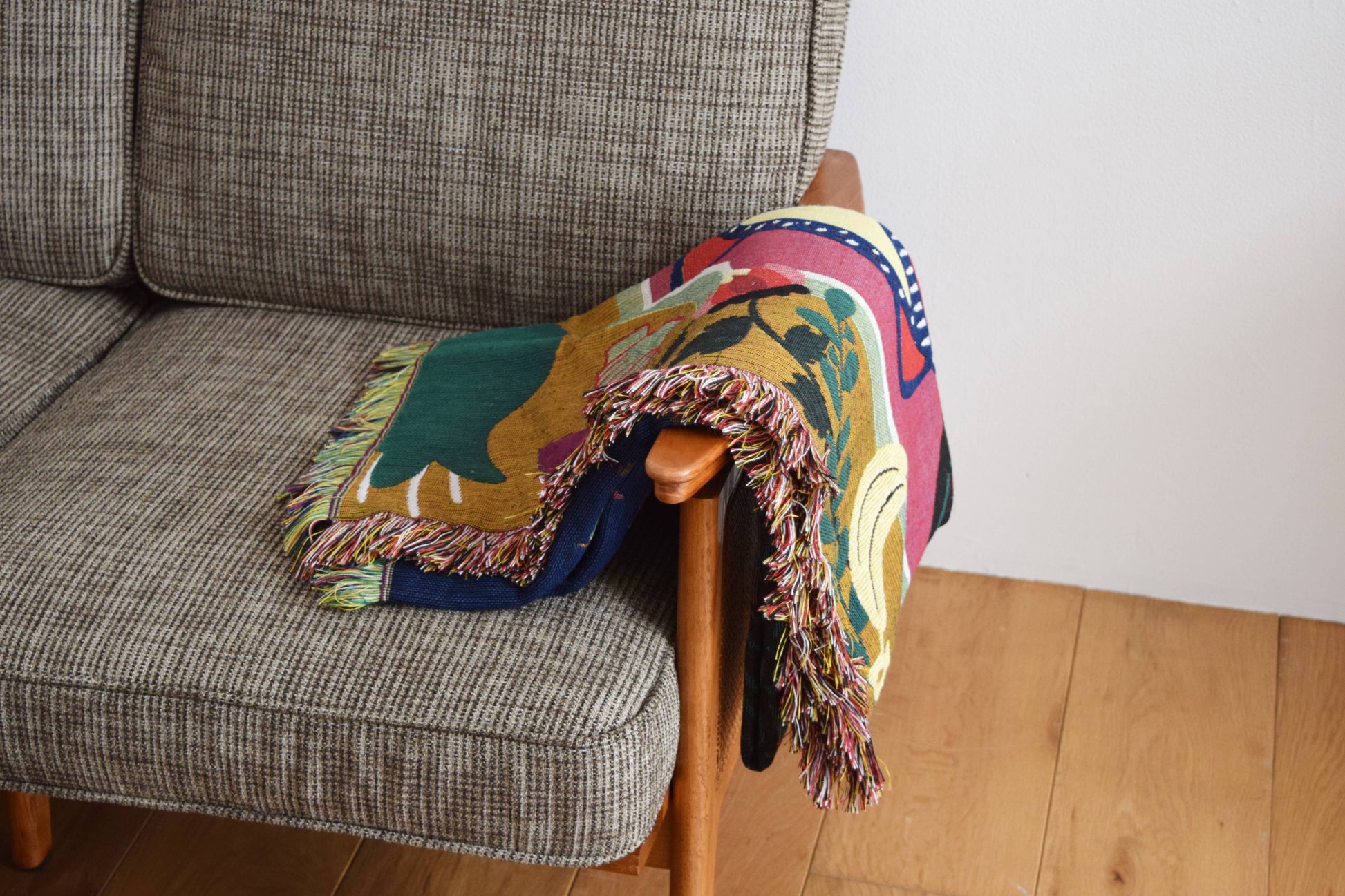 The DETAIL Gorilla Oax Throwket by DETAIL, a vibrant fringed multicolour blanket inspired by Oaxaca folk art, is draped over the wooden armrest of a gray fabric sofa on a light wooden floor beside a white wall.