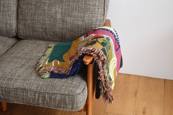 The DETAIL Gorilla Oax Throwket by DETAIL, a vibrant fringed multicolour blanket inspired by Oaxaca folk art, is draped over the wooden armrest of a gray fabric sofa on a light wooden floor beside a white wall.