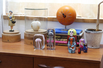 A wooden dresser displays the DETAIL Jellyfish Twisted Leg Square glass sculpture, painted nesting dolls, a woman figurine with flowers, a white apple, books, an orange clock, and a potted plant—adding eclectic style to modern décor.
