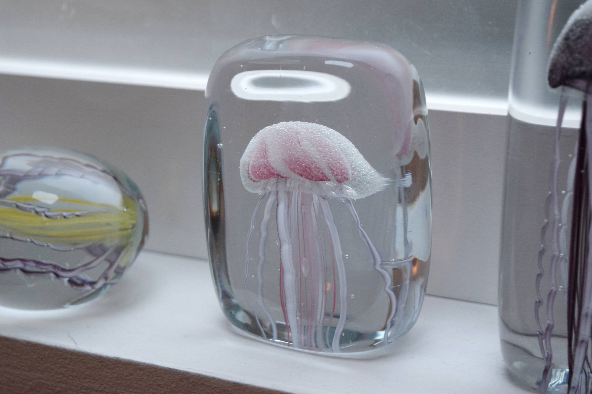 The DETAIL Jellyfish Twisted Leg Square by DETAIL, featuring an abstract pink and white jellyfish design, adds a modern touch to any space when displayed with other glass décor pieces.