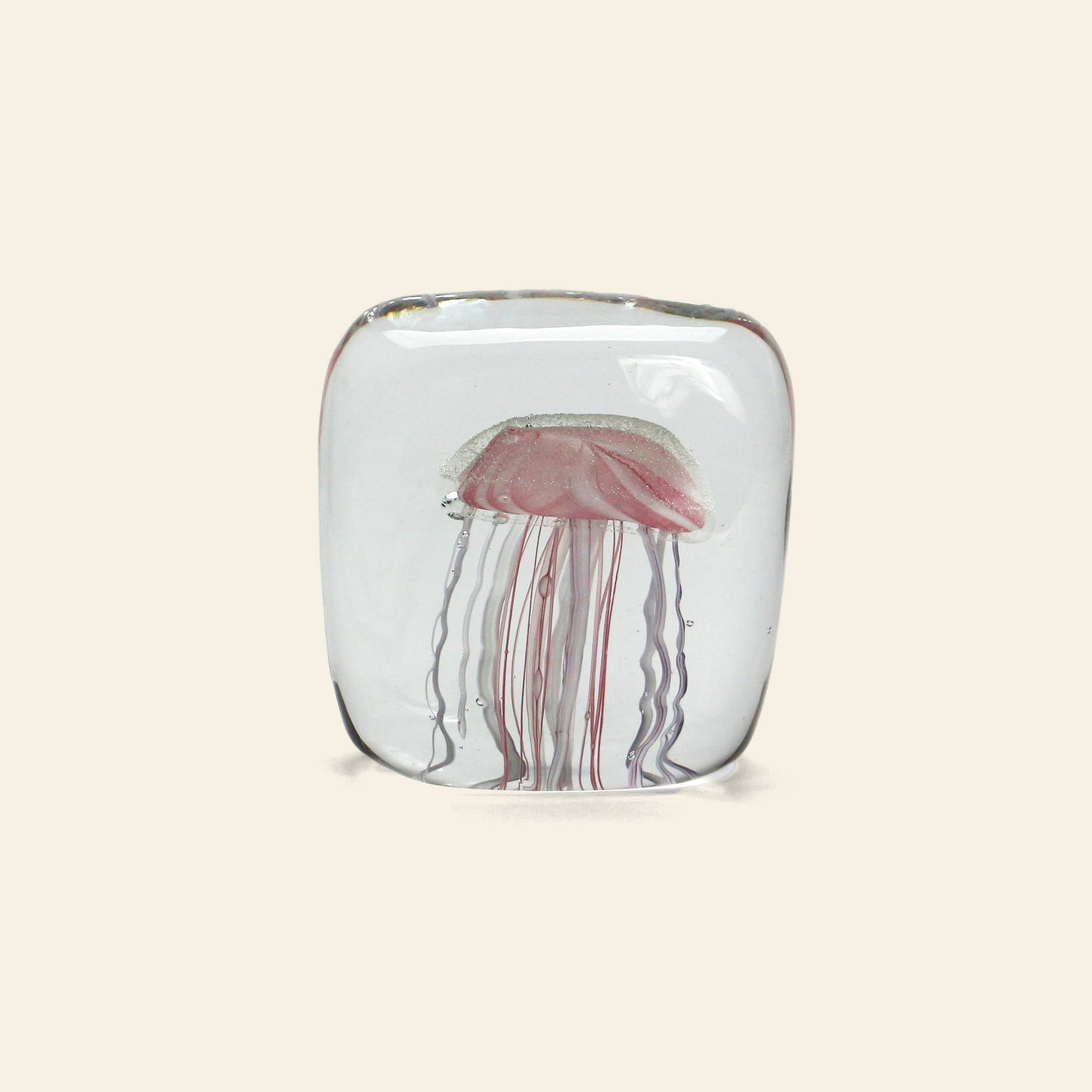The DETAIL Jellyfish Twisted Leg Square by DETAIL is a clear glass paperweight with a pink and white abstract jellyfish design, featuring long tendrils. This modern décor piece stands out against any light-colored background.