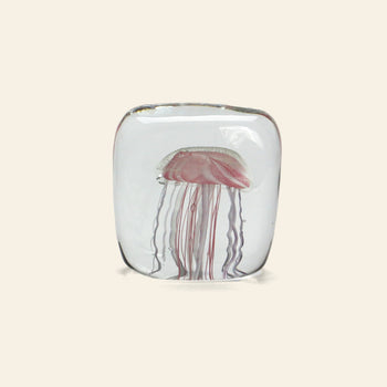The DETAIL Jellyfish Twisted Leg Square by DETAIL is a clear glass paperweight with a pink and white abstract jellyfish design, featuring long tendrils. This modern décor piece stands out against any light-colored background.