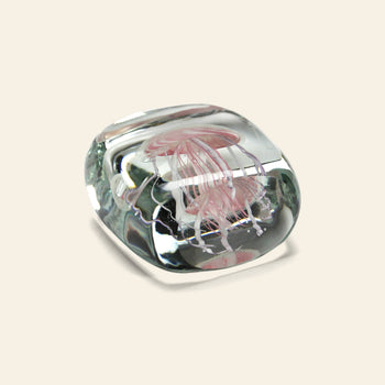 The DETAIL Jellyfish Twisted Leg Square by DETAIL is a clear glass paperweight featuring an abstract jellyfish design suspended inside, adding a modern décor touch against a light beige background.
