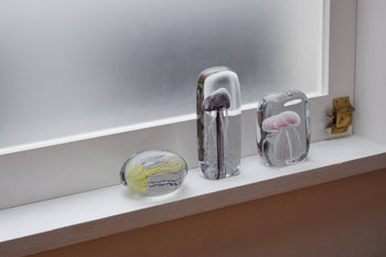 Three DETAIL Jellyfish Twisted Leg Square glass paperweights by DETAIL, featuring abstract jellyfish designs, sit on a white windowsill in front of a frosted window with a brass latch, adding modern décor to the scene.