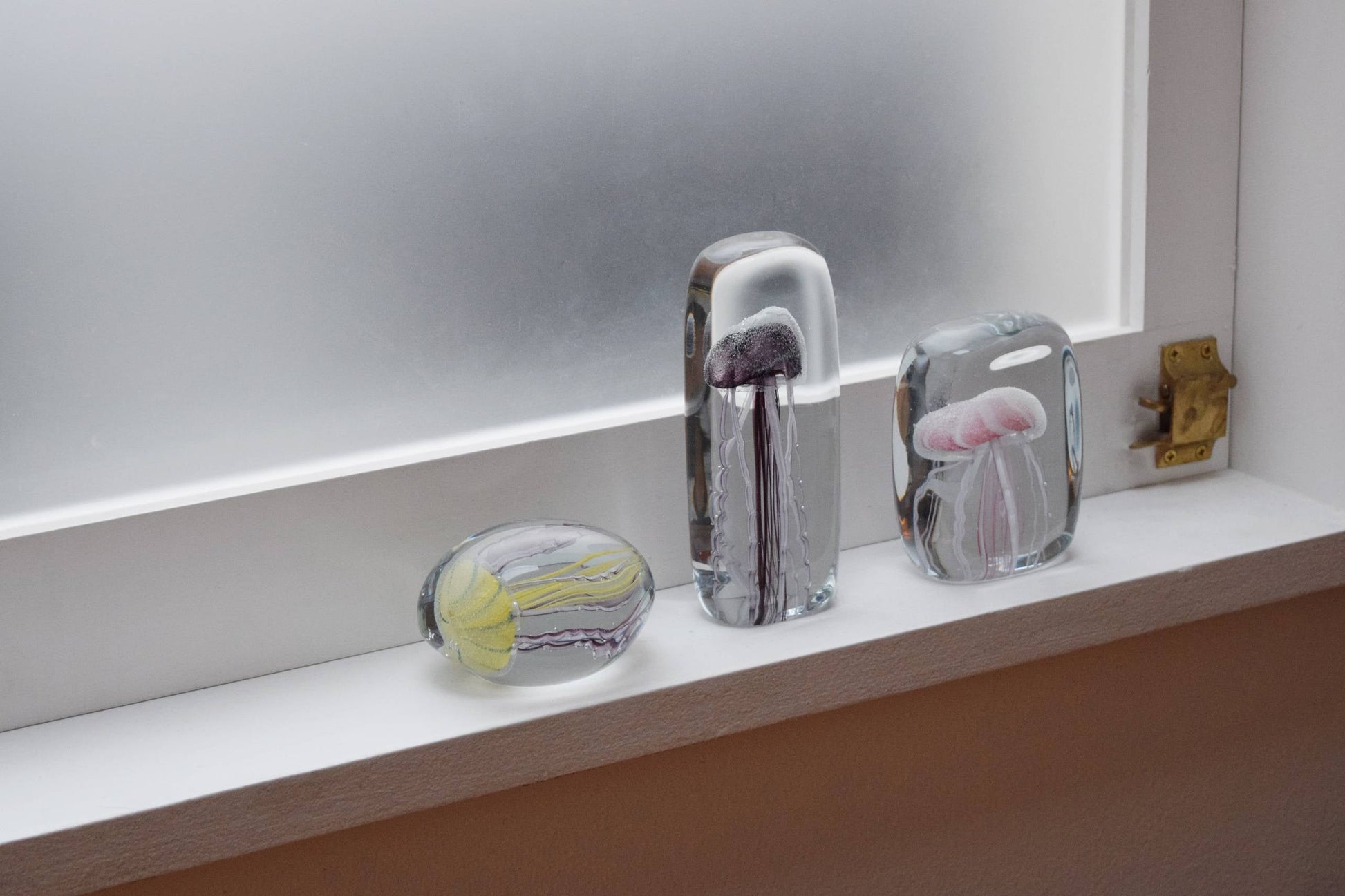 Three DETAIL Jellyfish Twisted Leg Tall glass paperweights—yellow, black, and pink—by DETAIL are displayed on a white windowsill before a frosted window, creating a striking modern art display.