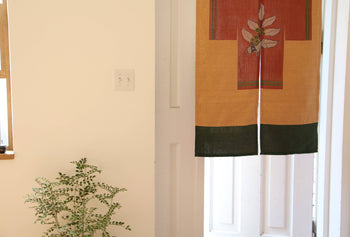 A small leafy green plant sits by a white wall and window. Nearby, a doorway features the DETAIL Noren Curtain from DETAIL, decorated with a plant-inspired yellow and green floral design.