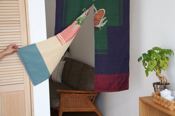 A hand pulls back the DETAIL Noren Curtain by DETAIL, revealing a cozy living room with a wooden chair, gray cushion, and a green leafy plant atop a small wooden cabinet.