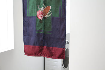 The DETAIL Noren Curtain by DETAIL, in dark blue and maroon with a mushroom and fern pattern, partially covers a white washing machine in a bright, minimalist room for a subtle, plant-inspired accent.
